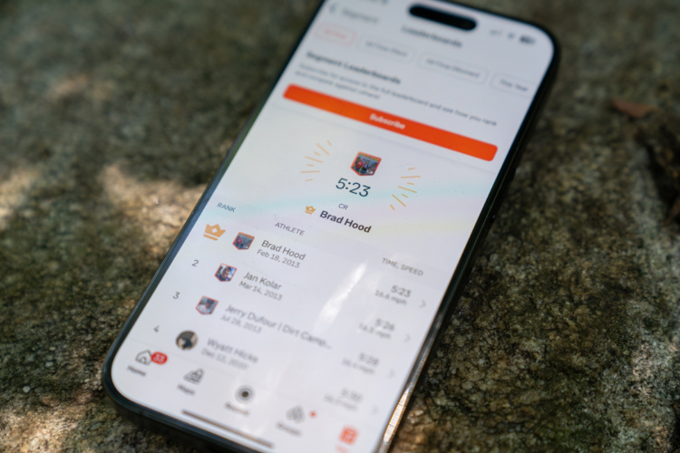 A close-up of a mobile phone displaying a leaderboard screen, featuring a ranking for the athlete Brad Hood with a record time of 5:23. The screen includes positions for other athletes and their respective recorded times. The phone is placed on a textured stone surface.