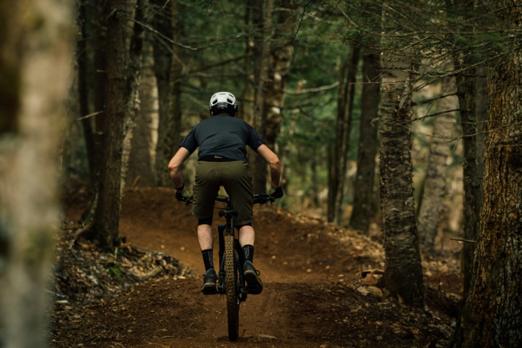 A person riding a mountain bike on a dirt trail through a forest, seen from behind. The rider is wearing a helmet, a black shirt, and brown shorts, and is navigating a curved section of the trail surrounded by trees. The scene is filled with lush greenery typical of a forest environment.