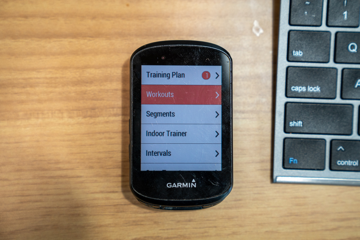 A top-down view of a Garmin device displaying a menu screen with options such as "Training Plan," "Workouts," "Segments," "Indoor Trainer," and "Intervals." The device is positioned on a wooden surface next to a computer keyboard.