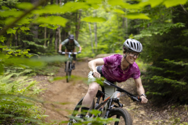 A mountain biker with a smile rides on a dirt trail surrounded by lush greenery, while another biker can be seen in the background navigating the path. The scene captures the excitement of biking in nature. Mont-Sainte-Anne mountain bike trail.