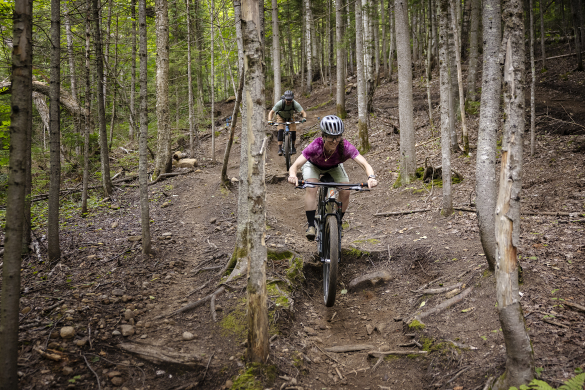 A woman in a purple shirt and shorts rides a mountain bike down a dirt trail in a wooded area, surrounded by tall trees. Another cyclist follows behind on the same path. The trail has patches of rocks and tree roots, highlighting the rugged terrain.