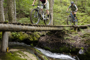 Two mountain bikers ride over a wooden bridge in a lush green forest. The first cyclist, wearing a helmet and a purple shirt, is in the foreground, navigating the bridge, while the second cyclist follows behind on a blue bike. Below the bridge, a gentle stream flows over moss-covered rocks. Mont-Sainte-Anne mountain bike trail.