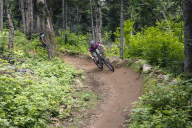 A mountain biker leans into a sharp turn on a dirt trail surrounded by dense greenery and trees. The scene captures the excitement of outdoor biking in a forested area. Mont-Sainte-Anne mountain bike trail.
