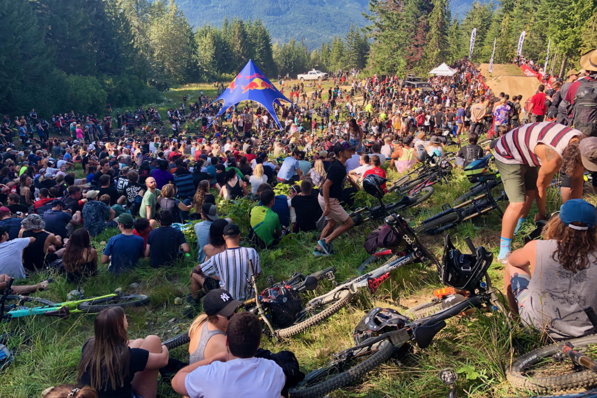 A large crowd gathers on a grassy hillside at Whistler surrounded by trees, with many people seated and some standing. Numerous mountain bikes are scattered throughout the scene, and a vibrant Red Bull tent is visible in the background, indicating an outdoor biking event or festival. The atmosphere is lively, with a mix of colorful attire and diverse participants enjoying the event under a clear blue sky.