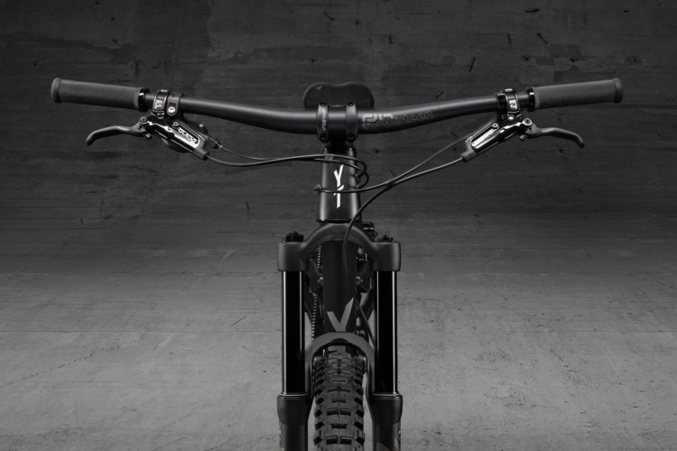 Alt text: Close-up view of a mountain bike handlebars and front suspension fork against a simple gray background. The handlebars feature black grips and brake levers, with cables neatly arranged.