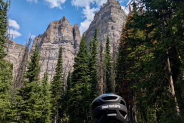 A person wearing a helmet and a patterned jacket stands on a hiking trail, gazing up at towering mountain peaks under a blue sky with scattered clouds. Surrounded by lush green trees and wildflowers, the scene captures a moment of adventure in a scenic outdoor landscape. CDT: Togwotee Pass to Jade Lake mountain bike trail.
