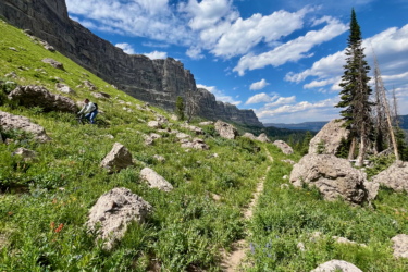 A scenic mountain landscape featuring a narrow trail winding through lush green grass and rocky terrain. Two people are sitting on rocks in the distance, surrounded by towering cliffs under a partly cloudy blue sky. The scene conveys a sense of tranquility and adventure in nature. CDT: Togwotee Pass to Jade Lake mountain bike trail.