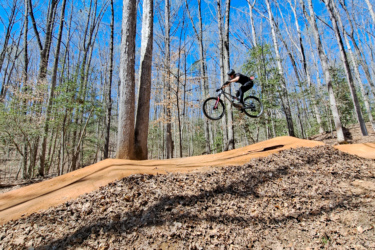 A mountain biker performing a jump on a dirt ramp in a wooded area, with trees in the background and a clear blue sky above. The ground is covered with leaves, and the biker is captured mid-air, showcasing a dynamic and adventurous moment. Pocahontas State Park mountain bike trail.