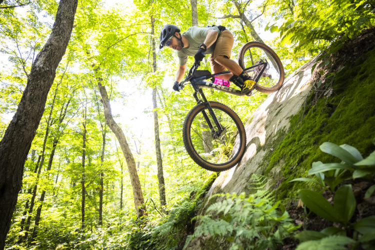 A mountain biker in a helmet rides down a rocky slope in a lush green forest, surrounded by tall trees and bright sunlight filtering through the leaves. The rider is focused and poised, showcasing skill and balance.
