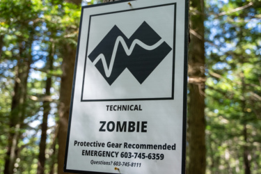 Sign indicating a "Technical Zombie" area, featuring a black and white graphic logo and text stating "Protective Gear Recommended," along with an emergency contact number and additional information, set against a backdrop of trees in a forest. Loon Mountain Resort mountain bike trail.