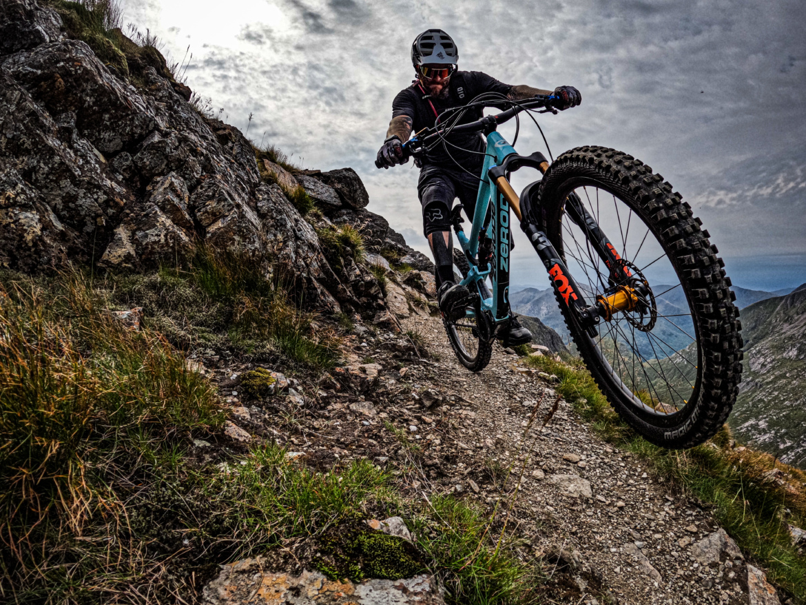 A mountain biker navigates a rugged trail with rocky terrain and grass, showcasing their skills as they lean forward on a blue and black mountain bike. The background features a scenic view of mountains under a cloudy sky.