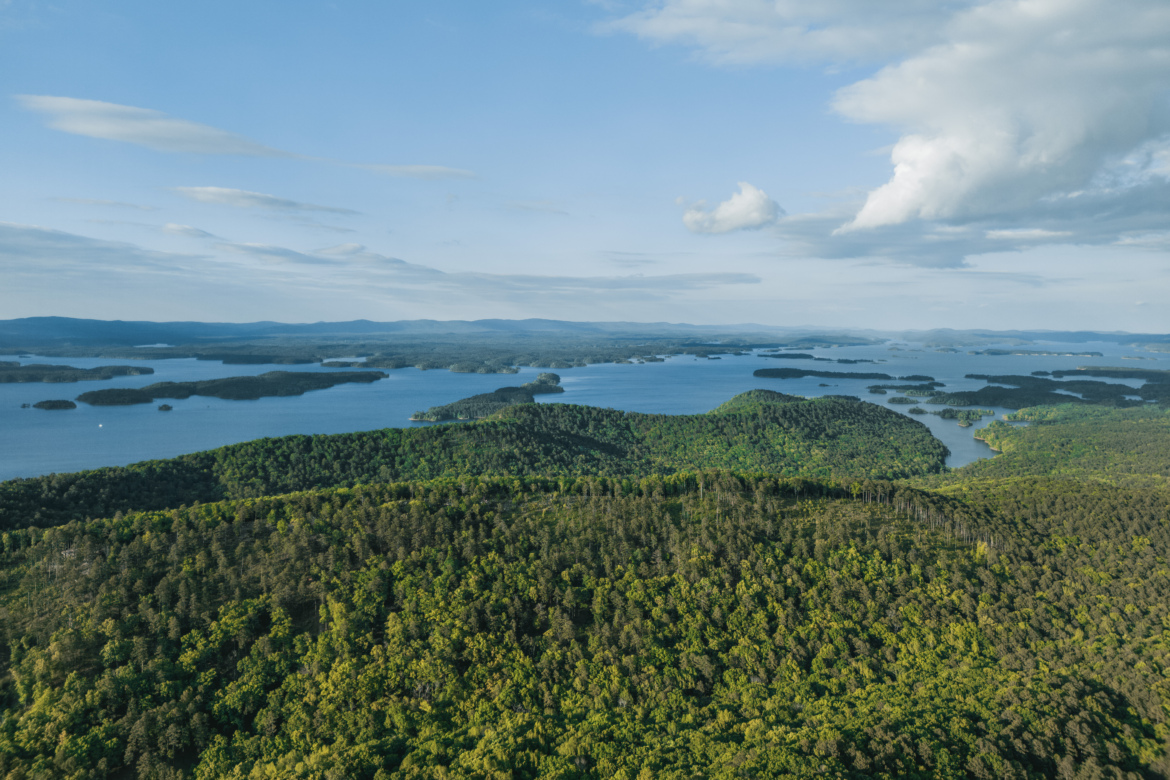 Aerial view of a lush green landscape with rolling hills and numerous blue lakes dotted throughout. The sky is clear with a few white clouds, and the horizon features distant mountains.