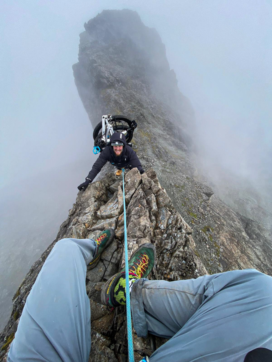 A climber sitting on a rocky ledge high in the mountains, wearing climbing gear and shoes, with a bicycle positioned behind them. A safety rope is visible connecting the climber to another person who is scaling the rock. The scene is shrouded in mist, and the rugged terrain emphasizes the height and exposure of the climb.