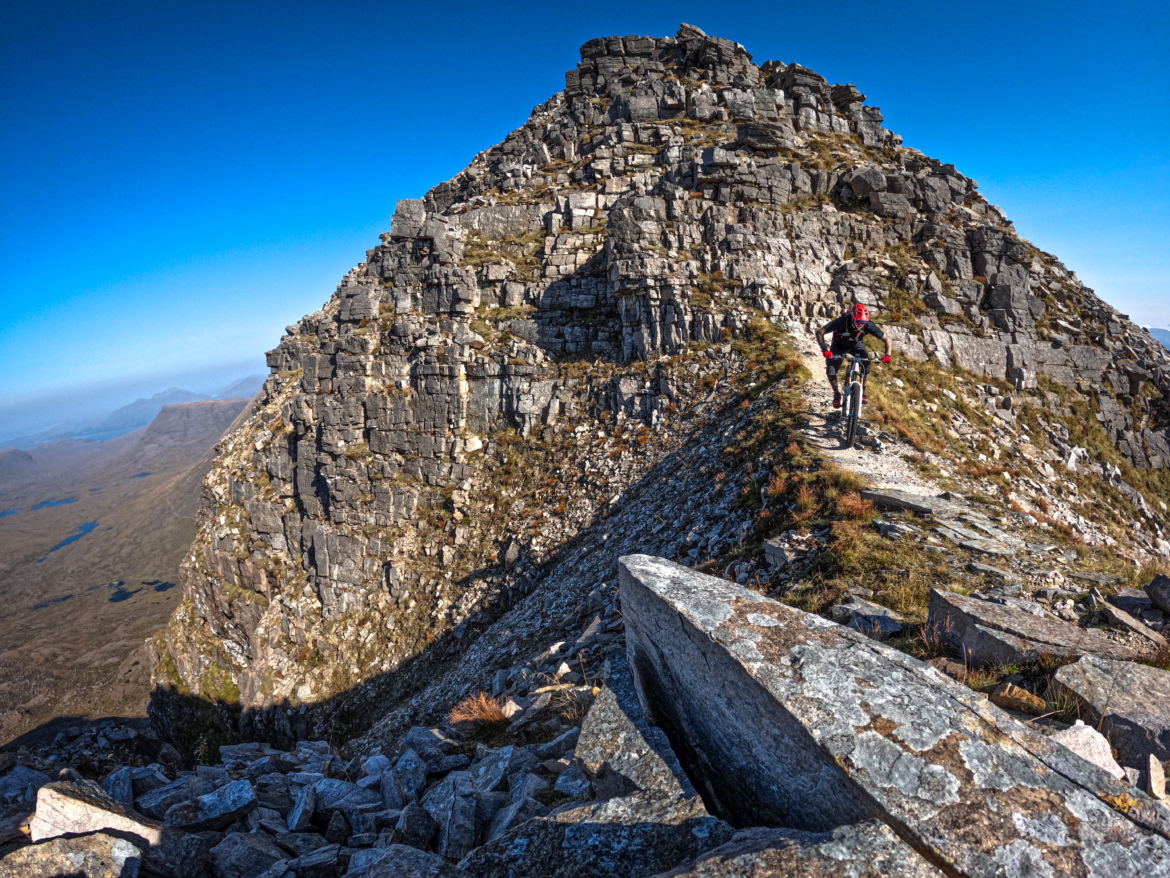A mountain biker navigates a narrow, rocky trail along a ridge with steep cliffs on either side. The landscape is rugged and mountainous, under a clear blue sky. In the background, rolling hills and lakes can be seen, showcasing the natural beauty of the area.
