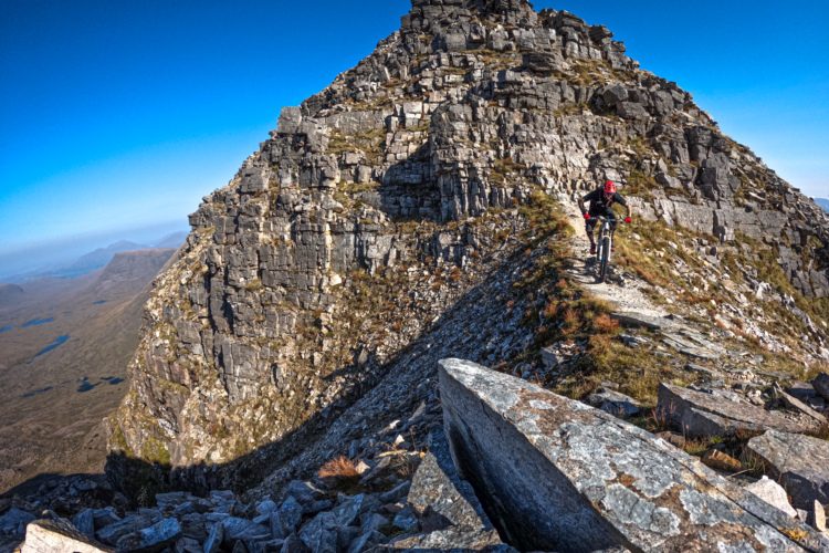 A mountain biker navigates a narrow, rocky trail along a ridge with steep cliffs on either side. The landscape is rugged and mountainous, under a clear blue sky. In the background, rolling hills and lakes can be seen, showcasing the natural beauty of the area.