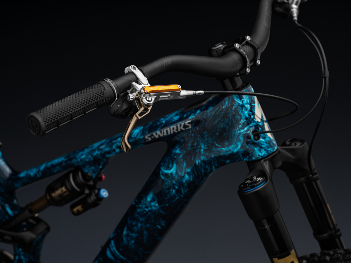 Close-up view of the handlebars and front fork of an S-Works mountain bike, showcasing a textured black grip and a gold hydraulic brake lever. The bike frame features a vibrant blue and black abstract design, with visible suspension components and a sleek, modern aesthetic.