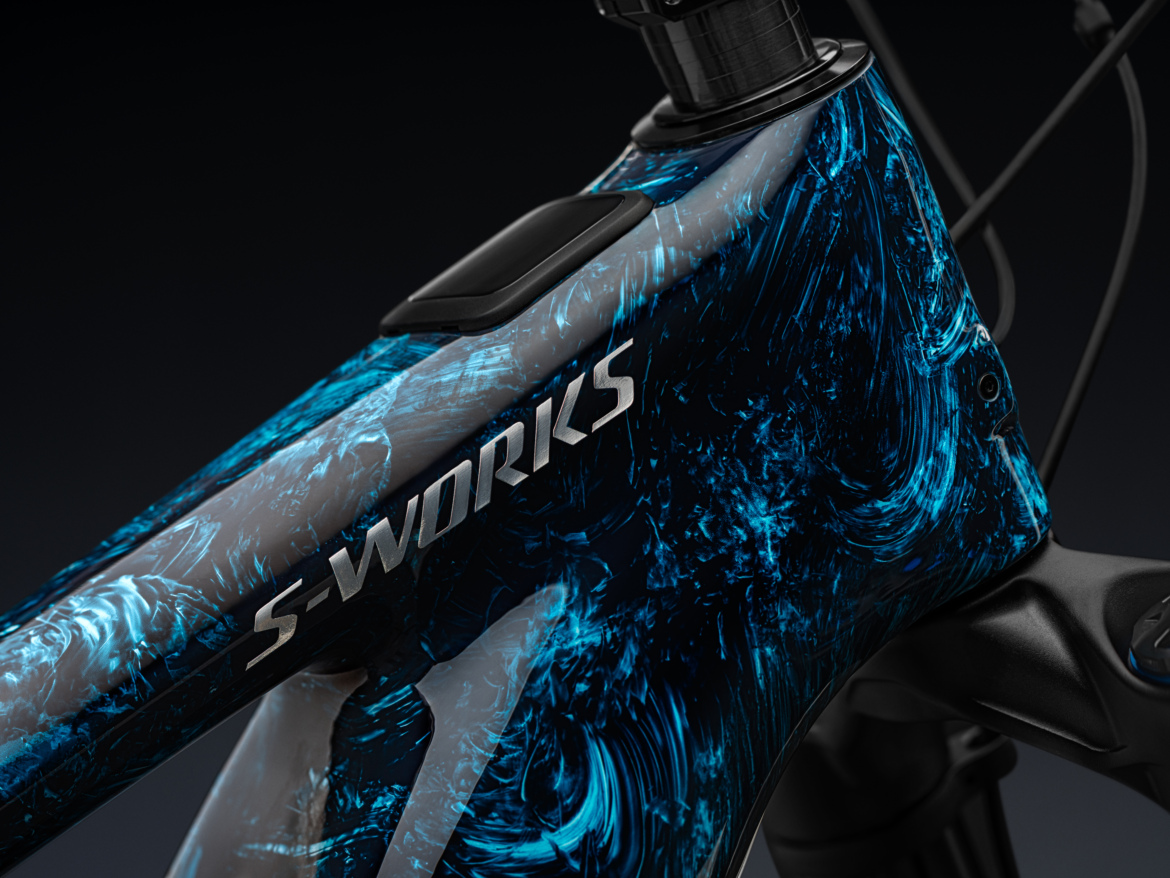 Close-up view of a mountain bike frame, featuring a vibrant metallic finish with swirling patterns in shades of blue and gray. The frame prominently displays the "S-Works" logo near the top tube, and the background is dark, emphasizing the bike's design and craftsmanship.