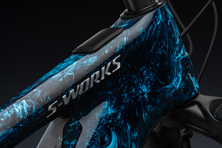 Close-up view of a mountain bike frame, featuring a vibrant metallic finish with swirling patterns in shades of blue and gray. The frame prominently displays the "S-Works" logo near the top tube, and the background is dark, emphasizing the bike's design and craftsmanship.
