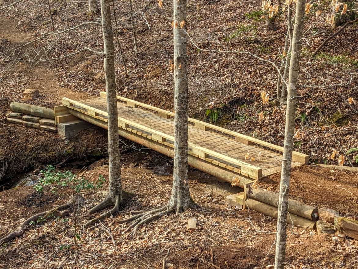 A newly constructed wooden bridge spans a small creek, surrounded by bare trees and a forest floor covered with fallen leaves. The bridge features a sturdy railing and is supported by wooden logs on either side. The path leading to the bridge is partially visible, indicating a natural trail through the wooded area.