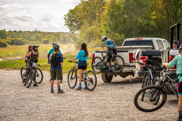 A group of mountain bikers in casual attire gather near a pickup truck parked on a gravel lot, preparing for a ride. Some riders are chatting, while others are loading bikes into the truck. The scene is set against a backdrop of trees and open fields under a partly cloudy sky. Wilderness Tours Bike Park mountain bike trail.