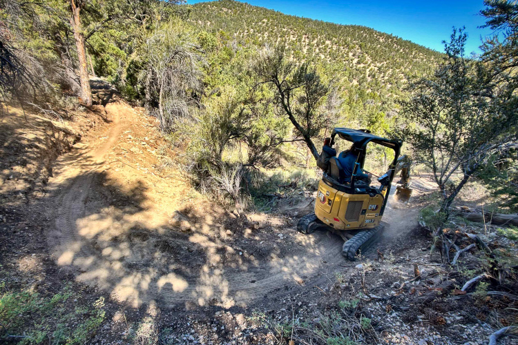 A yellow excavator maneuvering along a narrow, rocky trail in a forested area, surrounded by trees and hills under a clear blue sky. Dust is being kicked up as the machine makes its way along the path.