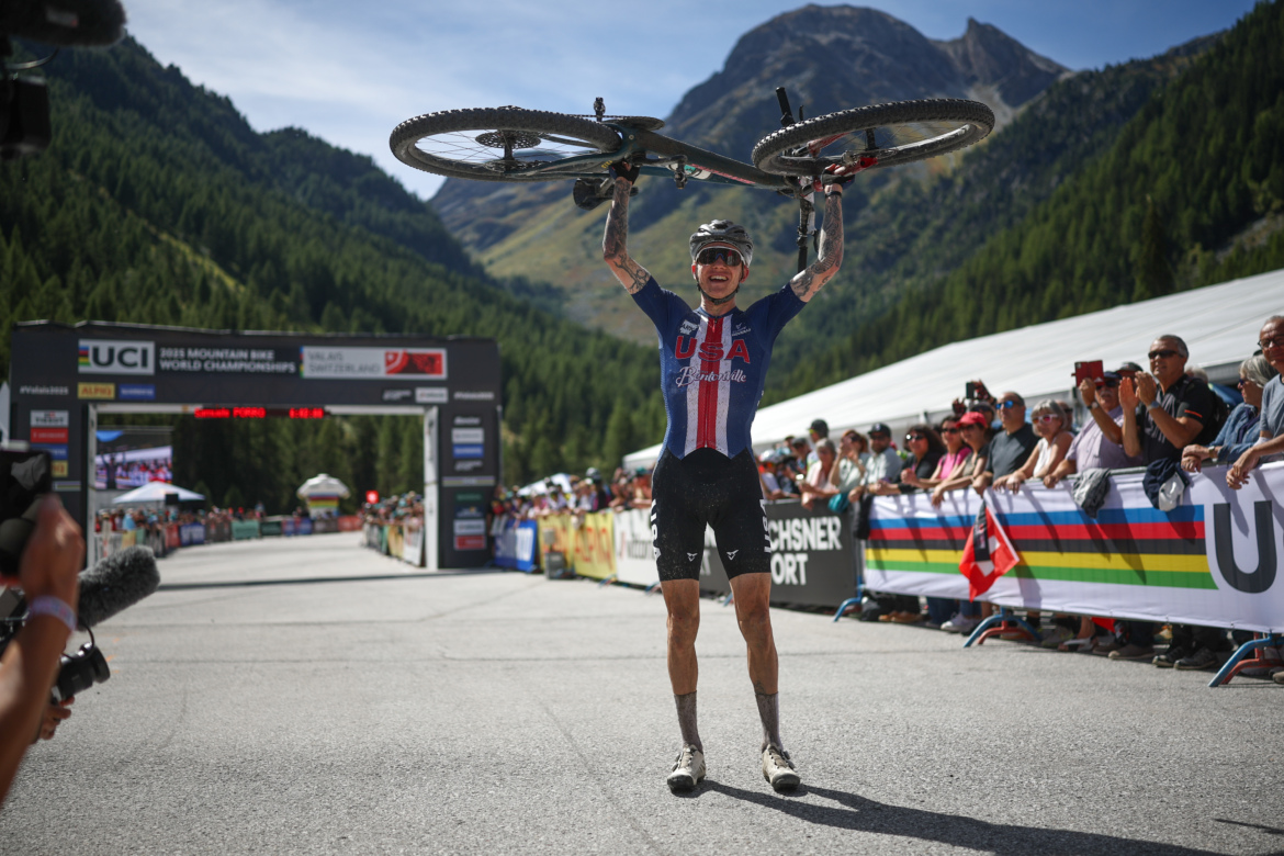 A mountain biker wearing a blue and red jersey with "USA" printed on it celebrates at the finish line, holding their bike overhead. The background features a mountainous landscape and an audience cheering. A banner for the 2023 UCI Mountain Bike World Championships is visible in the scene.