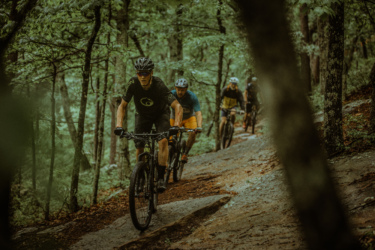 A group of four mountain bikers riding along a rugged forest trail, surrounded by lush green trees. The lead cyclist, wearing a black jersey and helmet, is navigating the rocky path, while the others follow closely behind. The scene captures a sense of adventure and camaraderie in a natural setting. Carvin's Cove Trail system mountain bike trail.