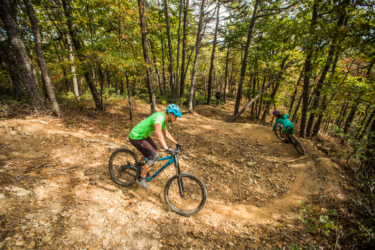 Mountain bikers navigate a rocky trail surrounded by trees in a forested area. Two riders are visible, one in a green shirt and blue helmet descending a slope, while another rider approaches a curve in the trail. The scene captures the dynamic movement and challenge of mountain biking in a natural setting. Carvin's Cove Trail system mountain bike trail.