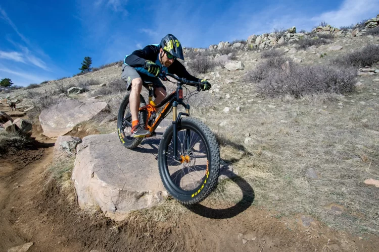 A mountain biker performing a jump off a large rock on a scenic dirt trail, surrounded by sparse vegetation and rocky terrain under a clear blue sky. The bike features wide tires, and the rider is wearing a helmet and athletic gear.