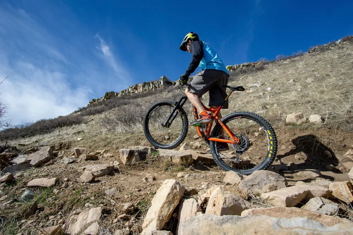 A mountain biker navigating a rocky trail, lifting the front wheel of their bike while riding uphill, set against a blue sky with wispy clouds and a grassy hillside in the background.