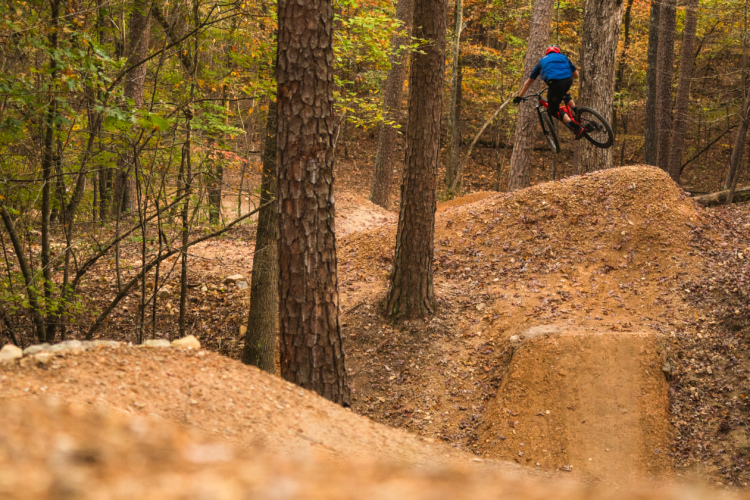 A mountain biker in a blue shirt and red helmet performs a jump on a dirt ramp in a forested area during autumn, with colorful leaves visible on the trees.
