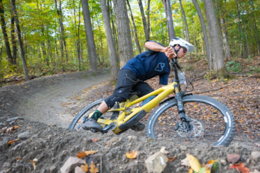 A mountain biker navigating a dirt trail in a forest, leaning into a turn while riding a yellow bike. The scene is set in autumn with trees showcasing colorful leaves, and the ground is covered with scattered leaves and dirt. Wilderness Tours Bike Park mountain bike trail.