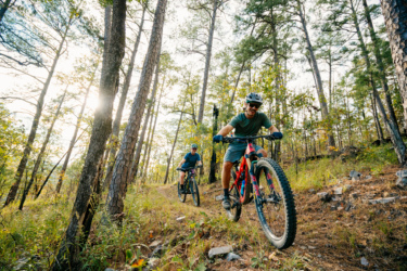 Two mountain bikers enjoying a trail ride through a lush forest with tall pine trees and dappled sunlight filtering through the foliage. One rider is smiling while maneuvering on a colorful bike, with the second rider following behind on a darker bike. The ground is covered with grass and scattered rocks, creating a natural outdoor setting. Lake Ouachita Vista Trail (LOViT) mountain bike trail.