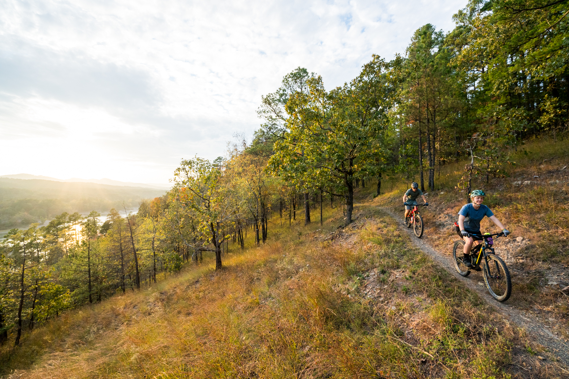 Two mountain bikers ride along a dirt trail through a forest with lush greenery, framed by trees. The sun is setting in the background, casting a warm glow over the landscape and illuminating the path ahead. The scene captures a sense of adventure and the enjoyment of outdoor activity. Lake Ouachita Vista Trail (LOViT) mountain bike trail.