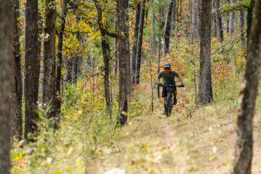 A cyclist riding a mountain bike along a narrow dirt trail through a dense forest, surrounded by trees with autumn foliage in shades of green, yellow, and red. Ouachita National Recreation Trail mountain bike trail.