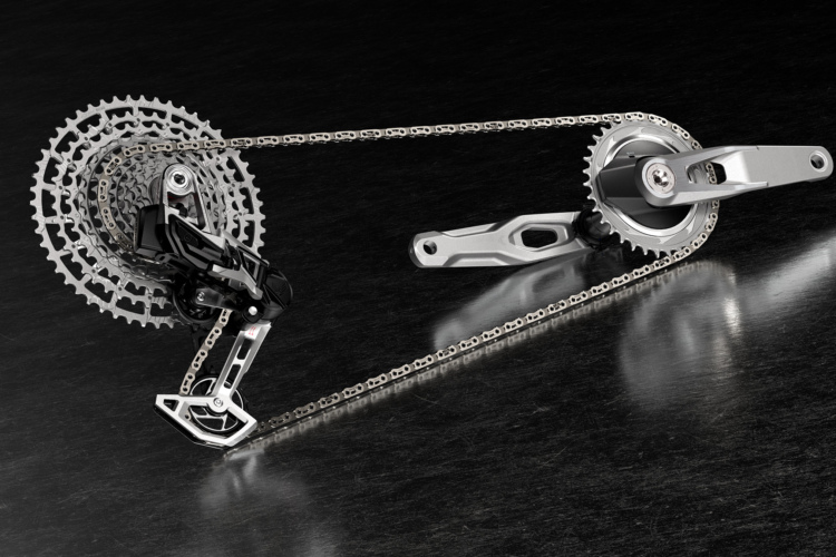 A close-up view of a bicycle drivetrain, showcasing a chain running over a silver cassette and crankset, featuring a black rear derailleur. The components are set against a dark, reflective surface, emphasizing their metallic finish and intricate design.