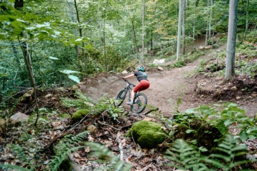 A mountain biker navigating a dirt trail through a lush green forest, with trees and ferns surrounding the path. The cyclist is leaning into a turn, wearing a helmet and colorful riding gear.