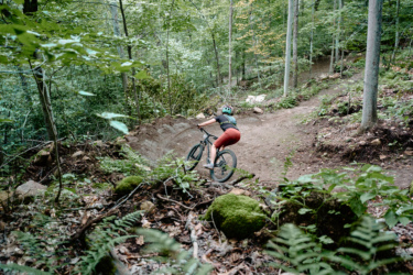 A person riding a mountain bike on a dirt trail through a lush green forest. The rider is navigating a curve in the trail, with trees and ferns visible in the background. Wilderness Tours Bike Park mountain bike trail.