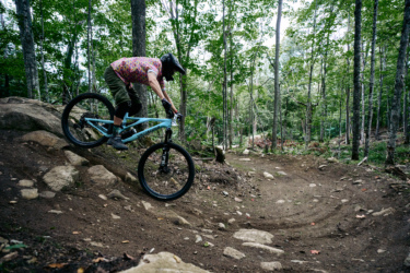 A mountain biker in a colorful shirt rides down a rocky trail in a forested area, surrounded by green trees and foliage. Wilderness Tours Bike Park mountain bike trail.