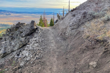 A narrow dirt trail winds along a rocky slope, with scattered stones and patches of dry grass on either side. In the background, a vast landscape of rolling hills and distant mountains is visible under a cloudy sky, creating a serene and natural setting. Beckwourth Peak Trail mountain bike trail.