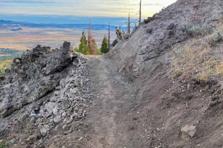 A winding dirt path leads along a rocky ridge, flanked by boulders and sparse vegetation. In the background, rolling hills and distant mountains stretch under a soft, cloudy sky, with hues of orange and yellow suggesting the onset of evening.