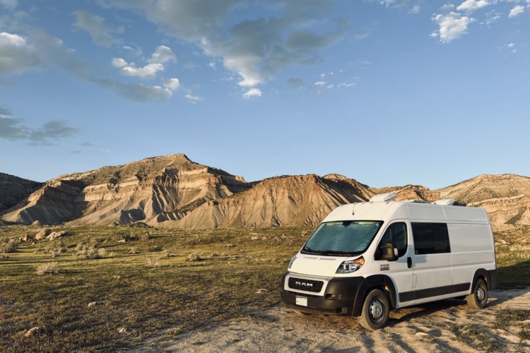 A white van parked on a dirt road in a scenic landscape featuring rugged mountains and a blue sky with clouds. The foreground shows grassy terrain with some rocky areas.