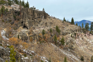 A rugged hillside featuring rocky outcrops and sparse vegetation, with a mix of evergreen and deciduous trees displaying autumn hues. The landscape is partly cloudy, revealing distant mountains in the background. Some areas show patches of snow, hinting at colder weather. Beckwourth Peak Trail mountain bike trail.
