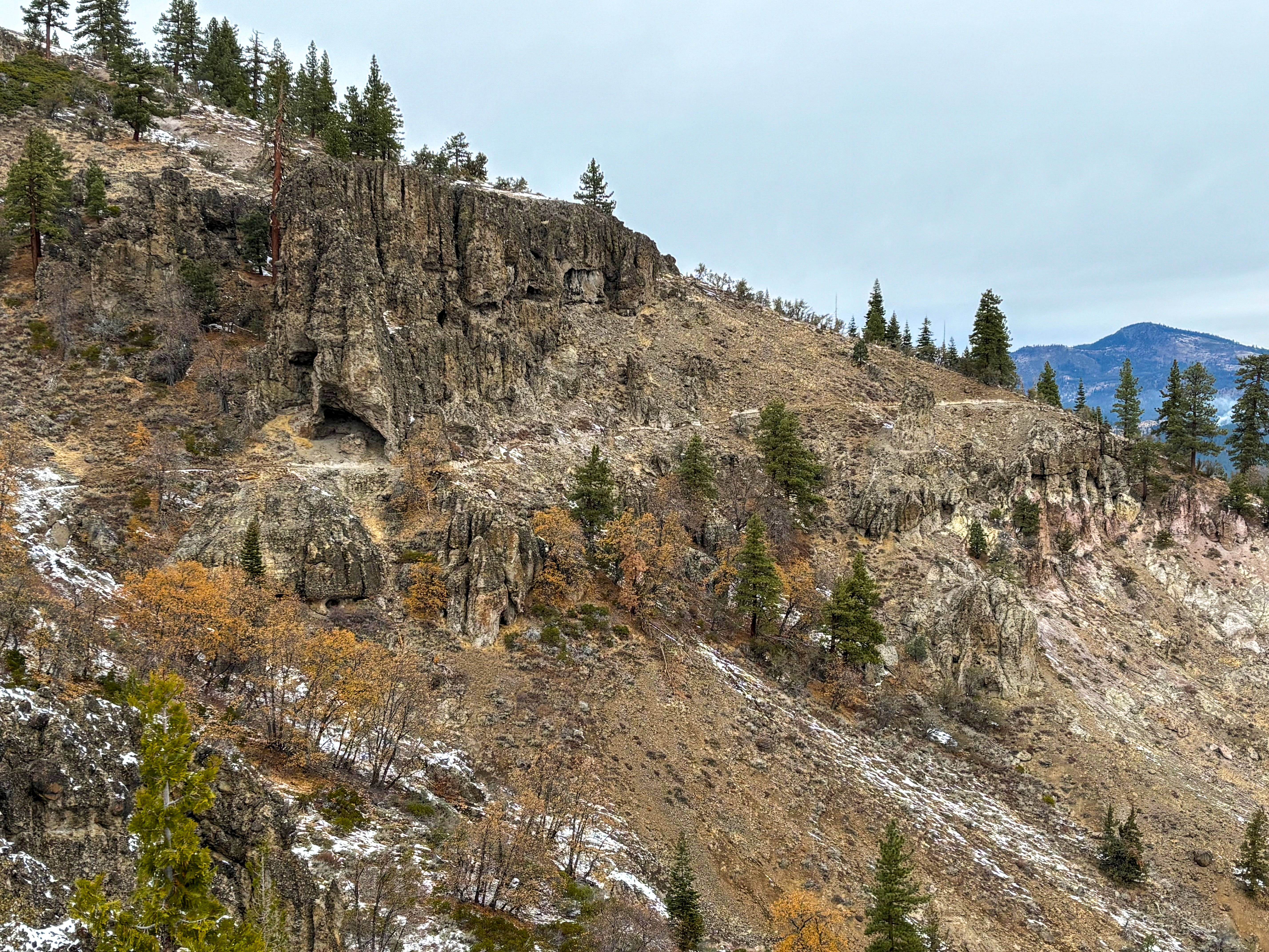 A rugged hillside featuring rocky outcrops and sparse vegetation, with a mix of evergreen and deciduous trees displaying autumn hues. The landscape is partly cloudy, revealing distant mountains in the background. Some areas show patches of snow, hinting at colder weather. Beckwourth Peak Trail mountain bike trail.