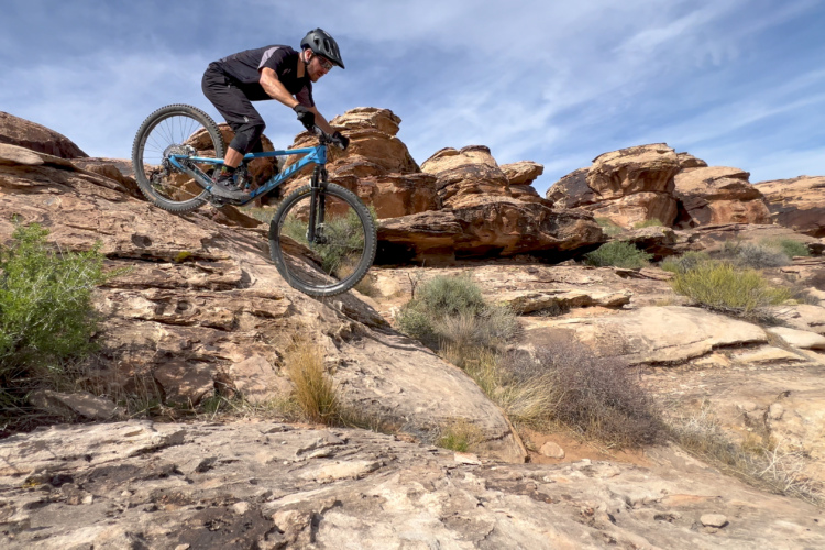 A mountain biker navigates a rocky terrain, descending from a rocky ledge on a blue bike. The landscape features large rock formations and sparse vegetation under a blue sky with scattered clouds.