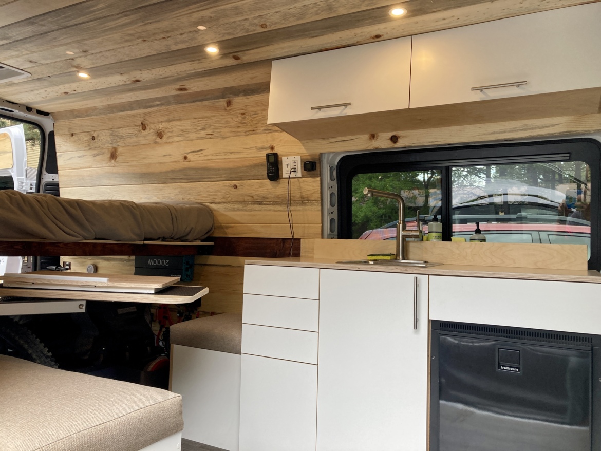 A spacious, modern van interior featuring wooden walls, a comfortable bed with a brown duvet, and a kitchenette area. The kitchen includes a sink, cabinetry, and a small refrigerator, with natural light coming through a side window. The overall design is sleek and functional, emphasizing a cozy, minimalist living space on the go.