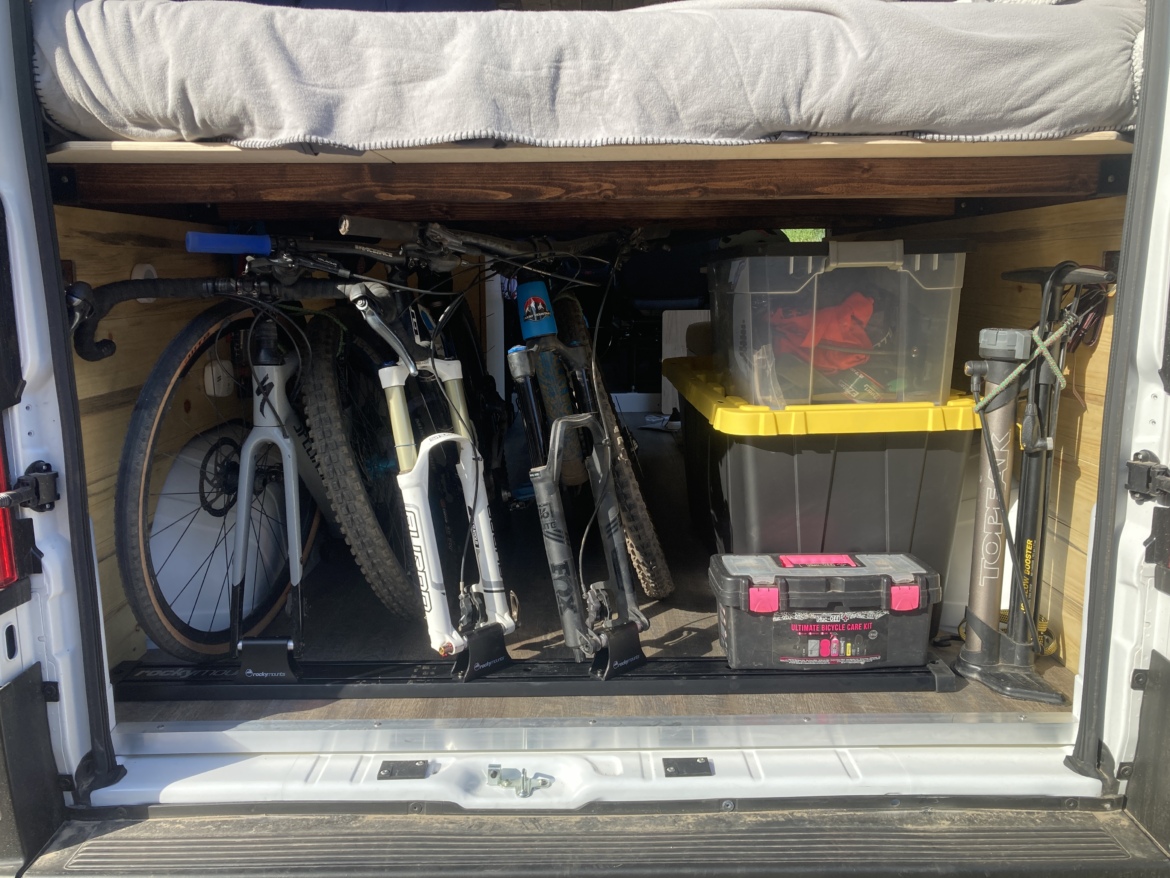 The interior of a van showcasing a well-organized storage space. Two mountain bikes are secured in bike stands, placed alongside plastic storage bins—one yellow and one black—filled with various items. A tool kit labeled "Ultimate Bicycle Care Kit" sits prominently on the floor, and a bed with gray bedding is visible at the top, creating a functional and cozy space for outdoor enthusiasts.