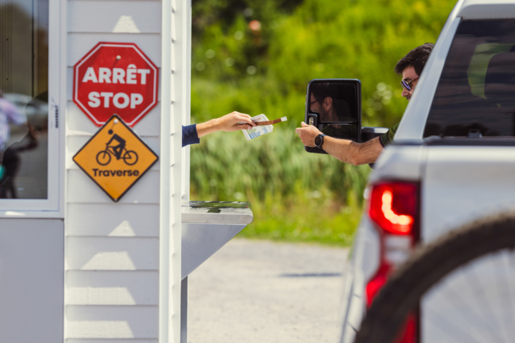 A vehicle at a checkpoint where a person inside the car is handing tickets to an attendant through a small window. Nearby signs indicate a stop and a bike crossing area, surrounded by greenery.