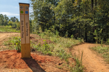 A wooden trail sign stands at a fork in the path, indicating directions to "Spirit Panda" to the right and "Bob Ross Pkwy" straight ahead. The sign features colored symbols representing the trail paths. The surrounding area is lush with greenery and trees, suggesting a natural outdoor setting. Saluda Confluence Recreation Area mountain bike trail.