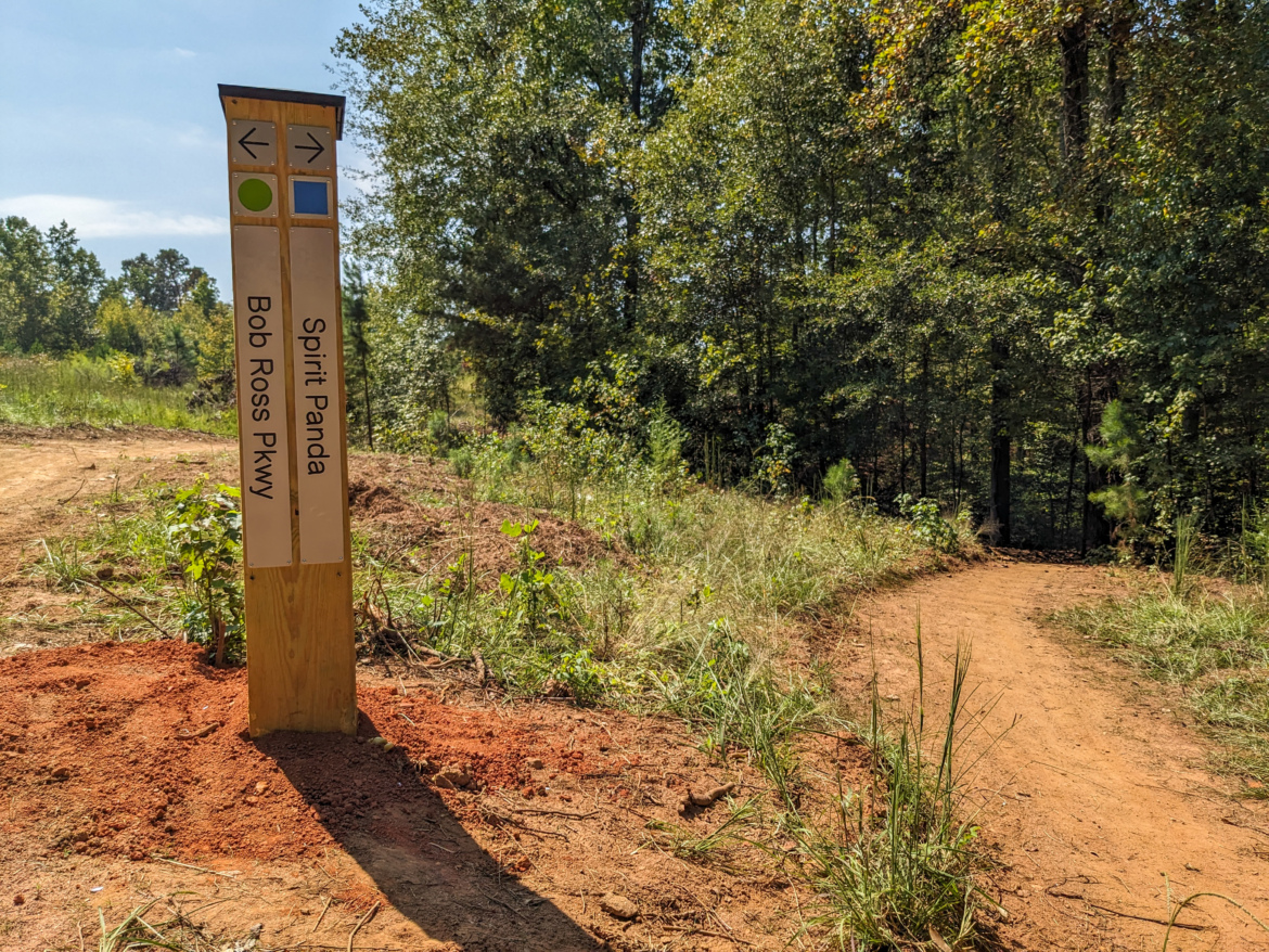 A wooden trail sign at a fork in the path, indicating directions for "Spirit Panda" and "Bob Ross Pkwy." The sign features arrows and colored markers, surrounded by lush greenery and sunlit trees in a natural setting. A dirt trail leads into the woods, flanked by grass and small plants.